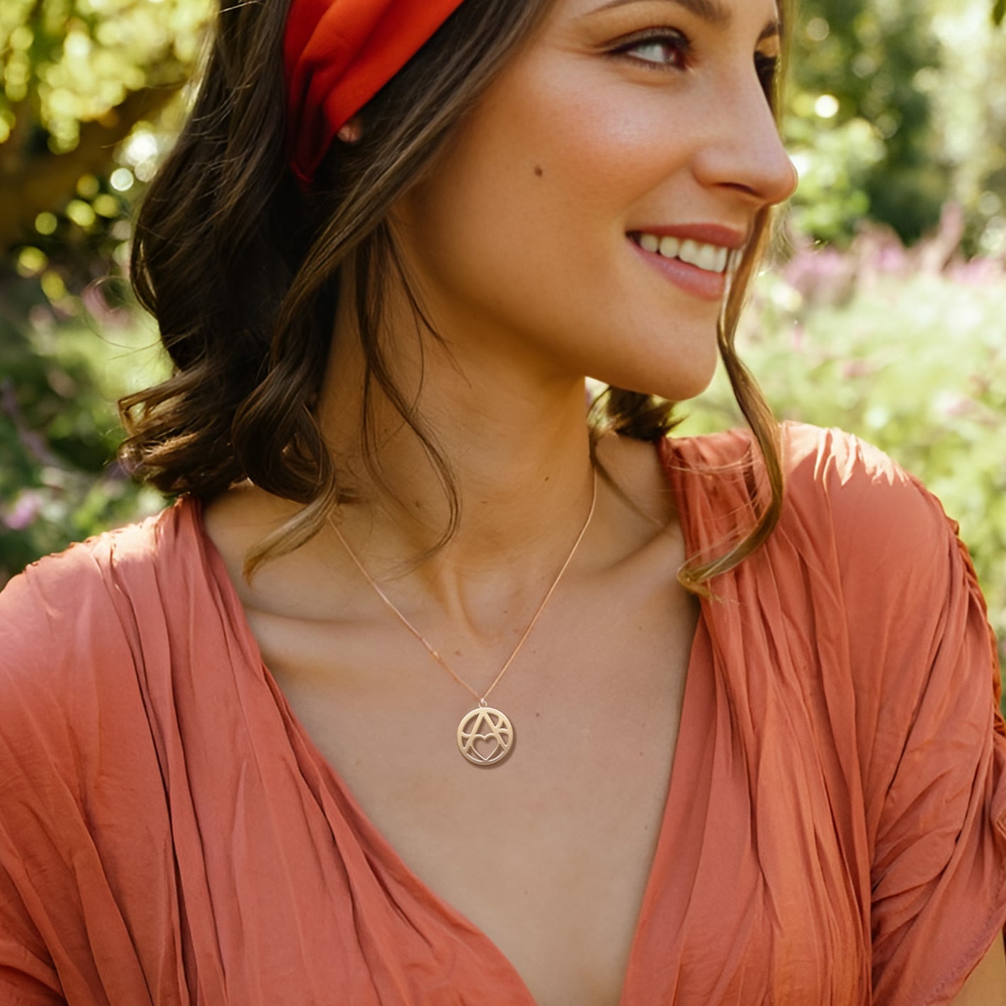 A smiling woman wearing a red headband and an orange dress wears a gold circular monogram pendant while standing in a sunny outdoor setting.