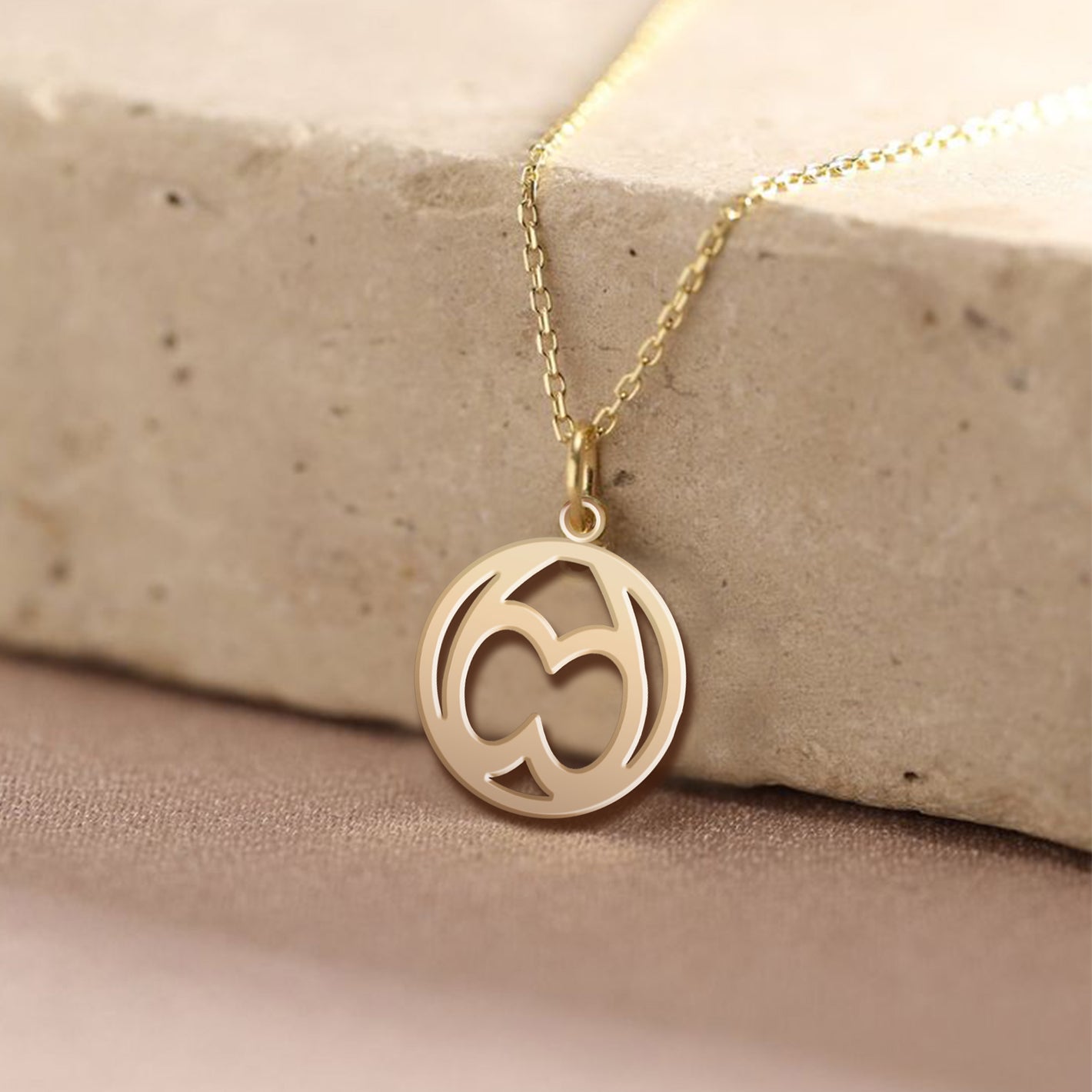 A gold circular pendant necklace with an abstract symmetrical monogram is displayed resting against a light-colored stone surface.