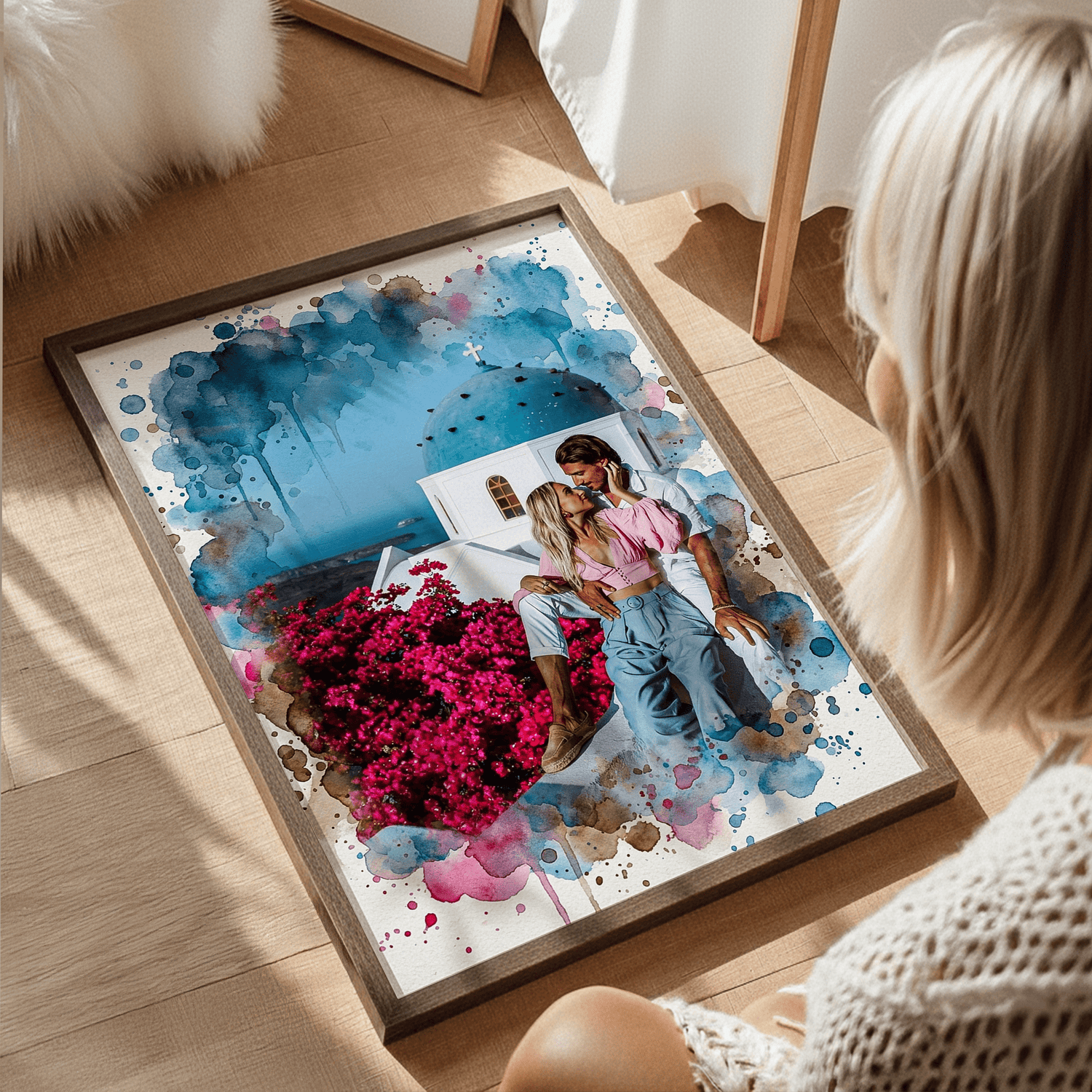 Woman viewing a framed personalized watercolor print of a couple kissing in Greece, surrounded by pink bougainvillea and blue and magenta paint splatters.