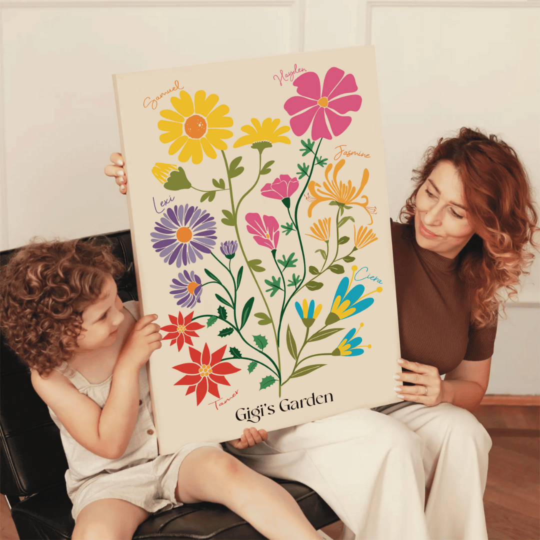 A woman and a young child with curly hair sit together on a black leather chair, holding a large "Gigi’s Garden" birth flower canvas.