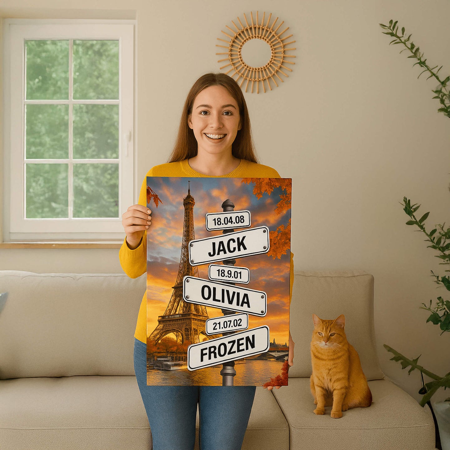 A woman in a yellow sweater sits on a beige sofa next to an orange cat, holding a personalized canvas of the Eiffel Tower featuring street signs with names and dates.