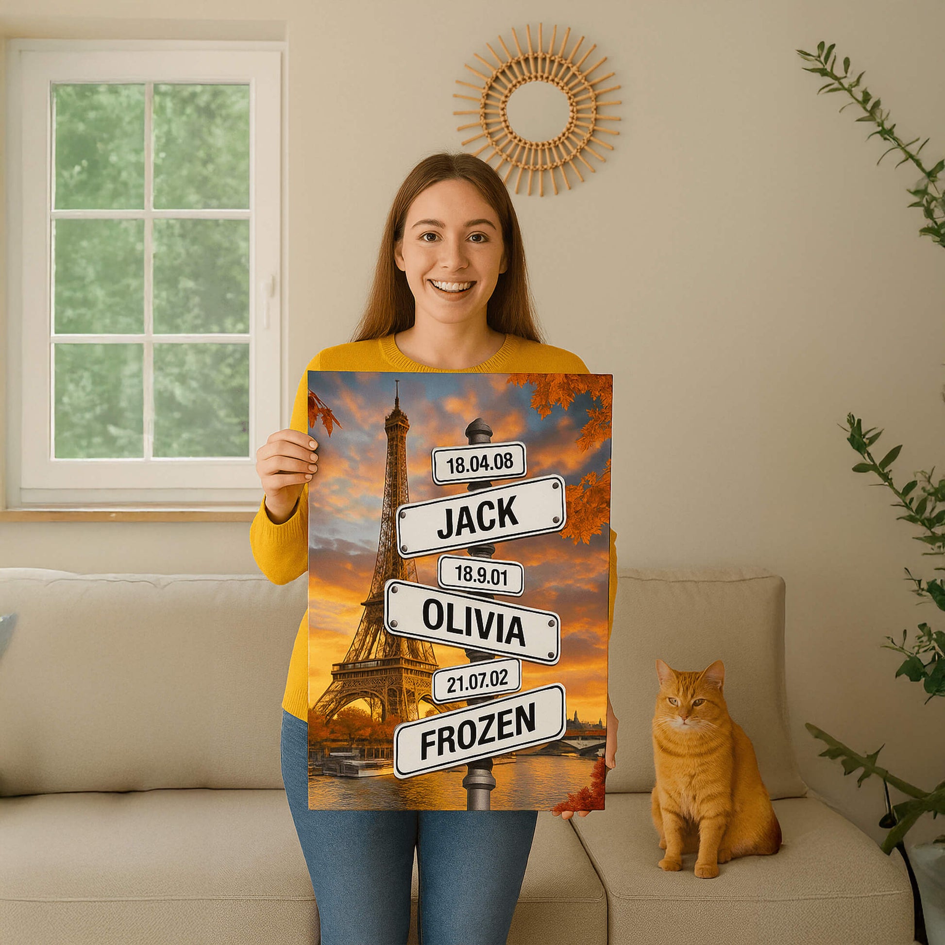 A woman in a yellow sweater sits on a beige sofa next to an orange cat, holding a personalized canvas of the Eiffel Tower featuring street signs with names and dates.