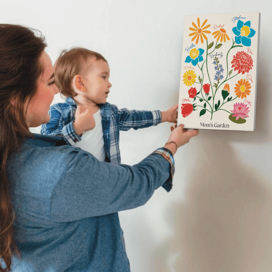 A mother and toddler hang a "Mom's Garden" birth flower canvas on a white wall.