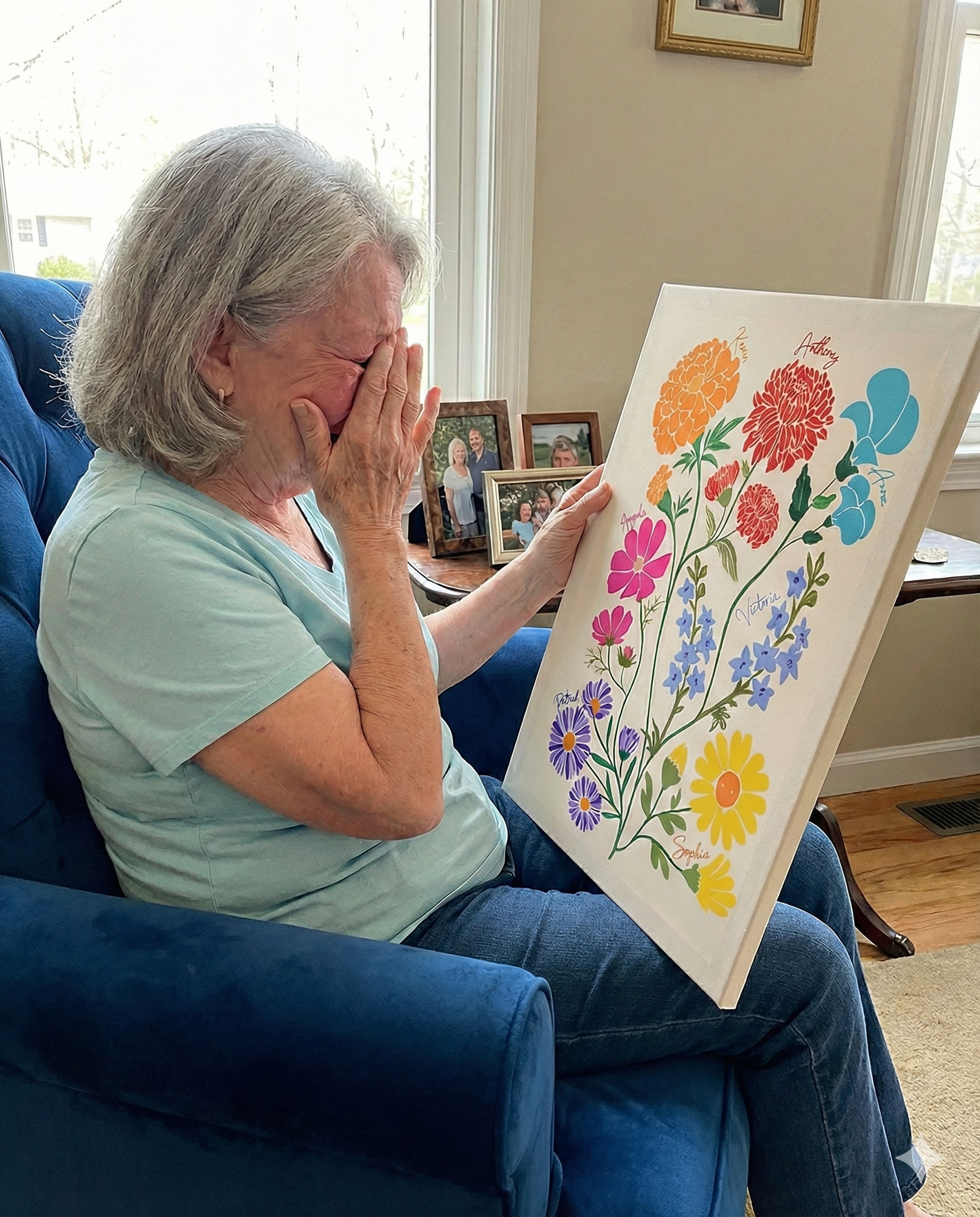 Older woman sitting in a blue chair, crying and covering her face, while holding a custom canvas print of colorful, labeled flowers.