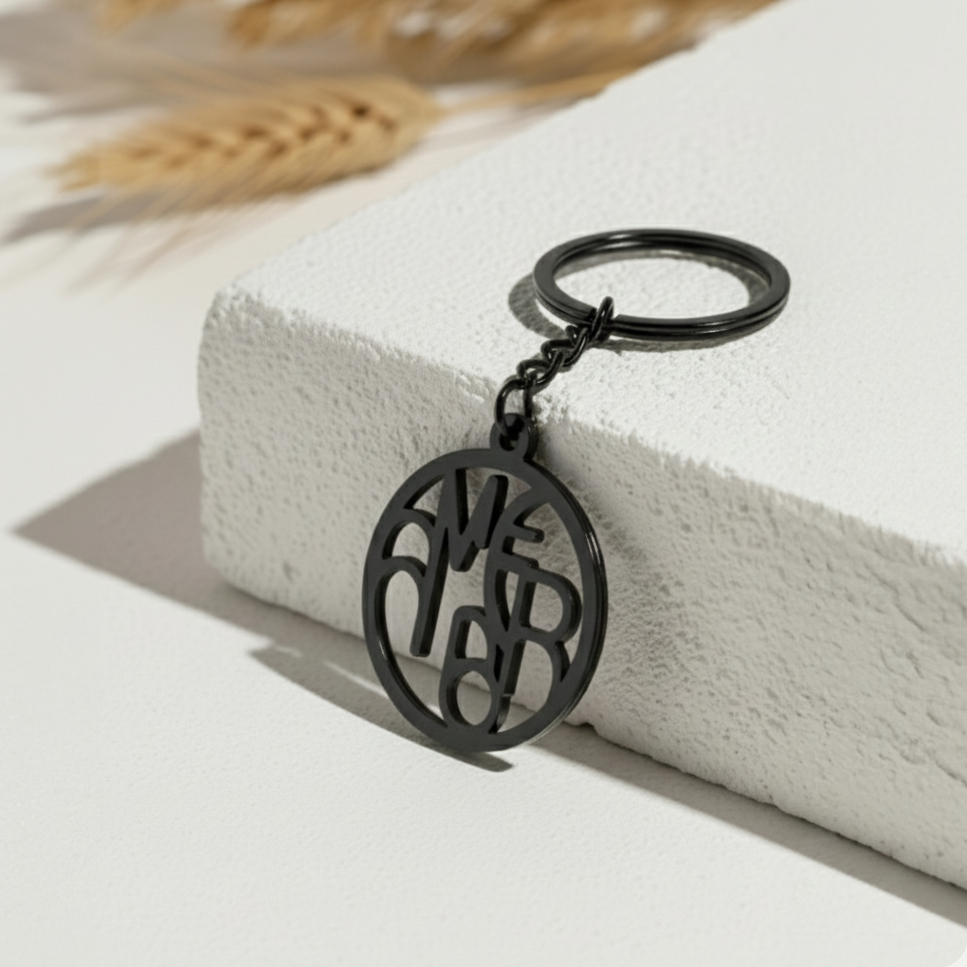 A modern black circular keychain with custom monogram lettering "ME" and "RB" cutouts. The keychain rests at an angle on a textured white stone surface, with soft natural shadows and blurred dried wheat stalks in the background.