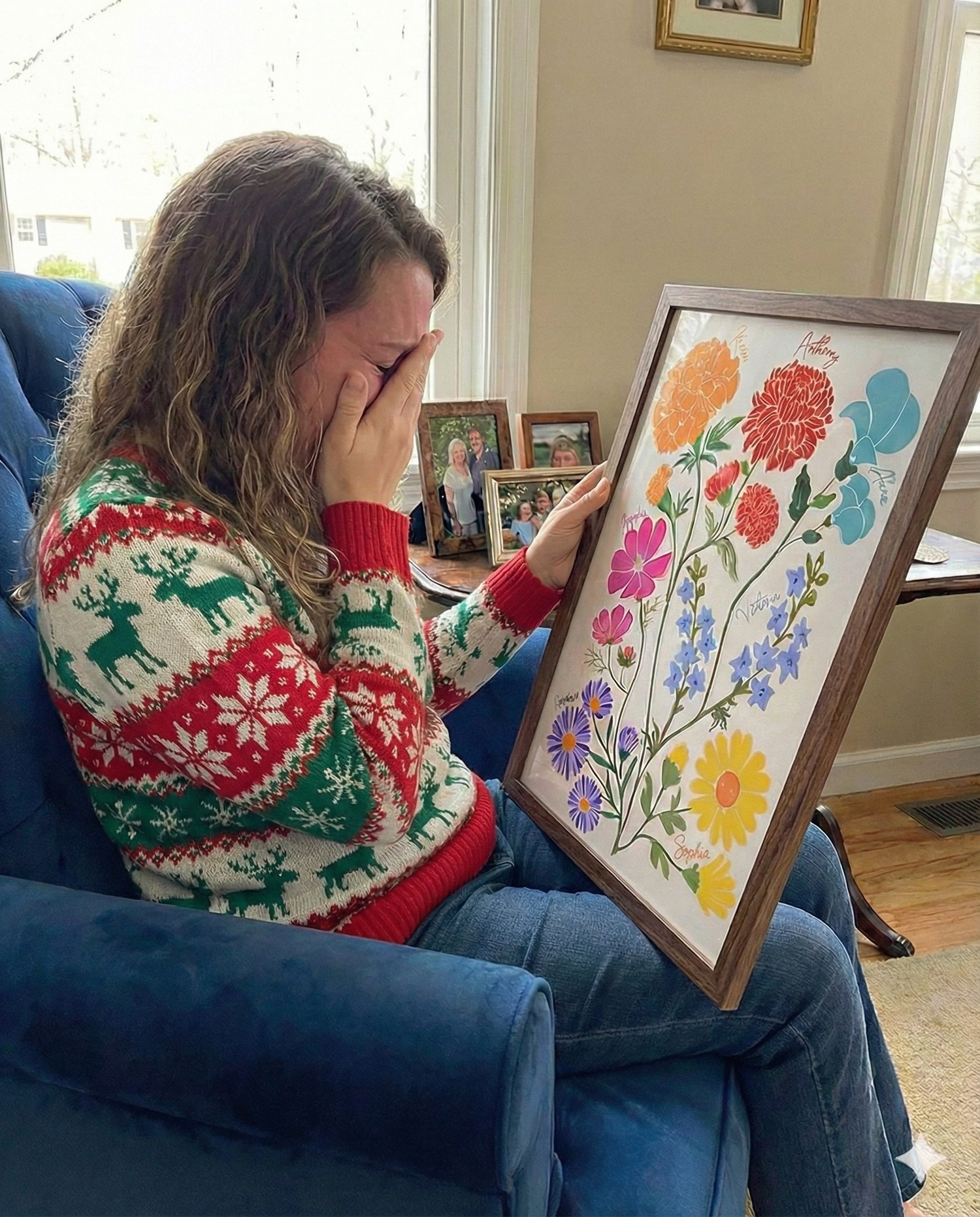 Woman in a Christmas sweater crying while holding a framed colorful custom garden print with labeled flowers.