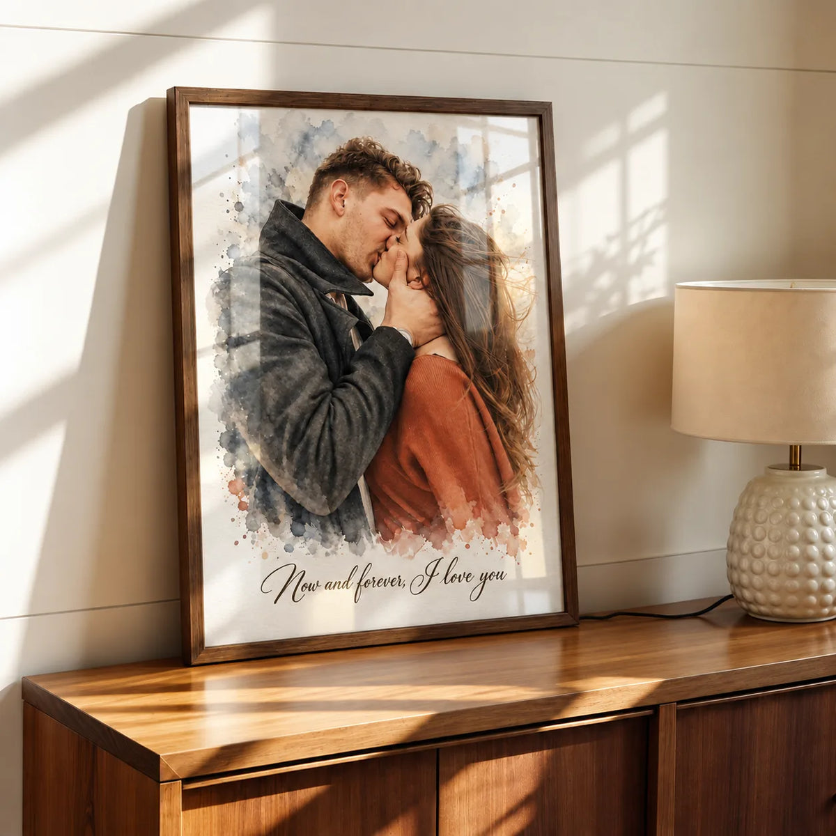A framed watercolor portrait of a couple kissing leans on a wooden sideboard.