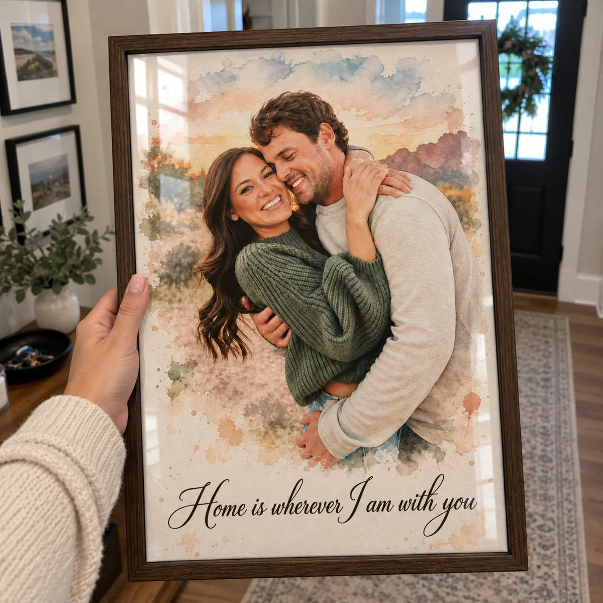 A hand holds a framed watercolor portrait of a couple hugging in a field at a front door with a wreath.