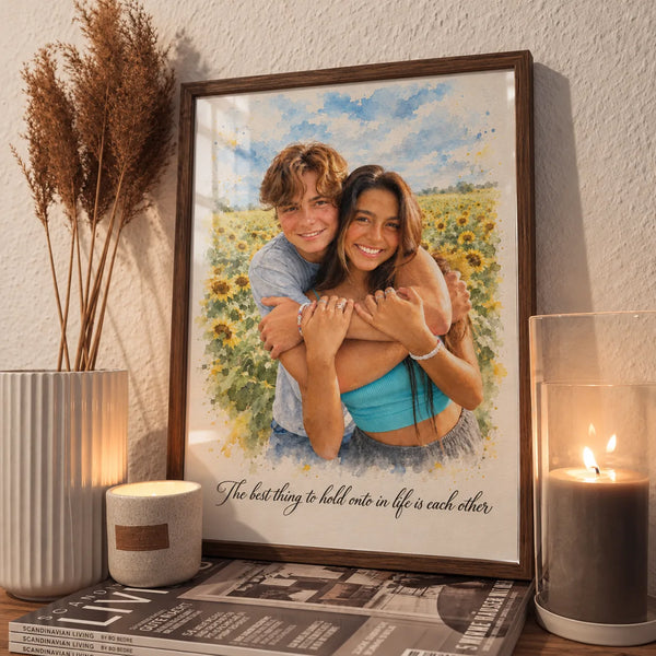 A framed watercolor portrait of a couple in a sunflower field leans beside a candle and dried grass.