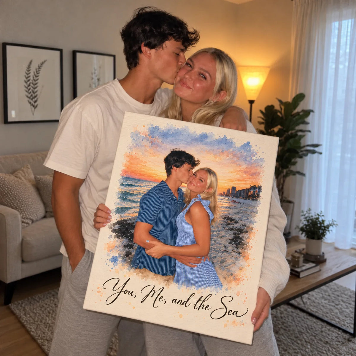 A couple stand close and hold a canvas watercolor portrait of themselves at the beach, he kissing her cheek.