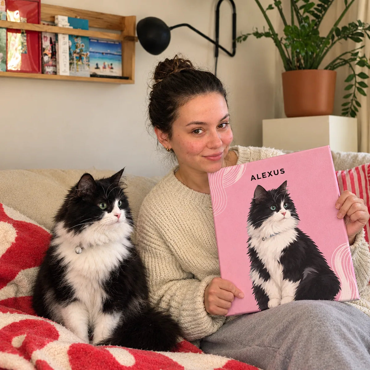 A smiling woman on a sofa holds a pink canvas portrait of her black and white cat while the real cat sits next to her.