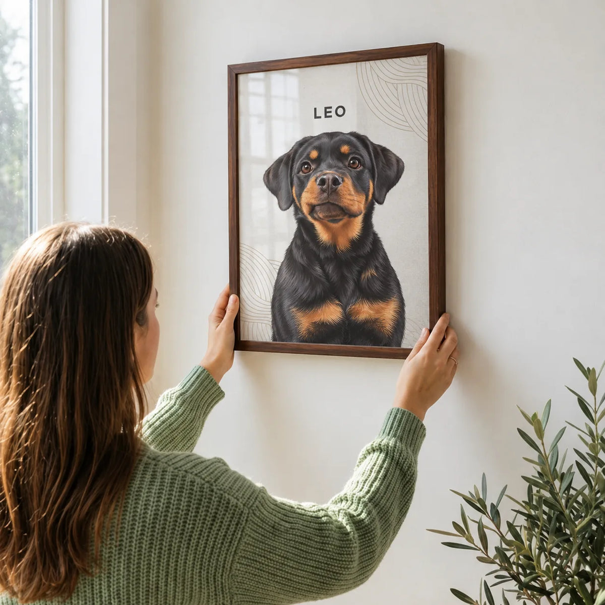 A woman in a green sweater hangs a framed portrait of a Rottweiler on a white wall.