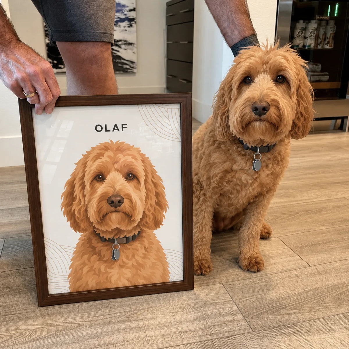 A person holds a framed portrait of a goldendoodle while the real dog sits beside it on a wood floor.